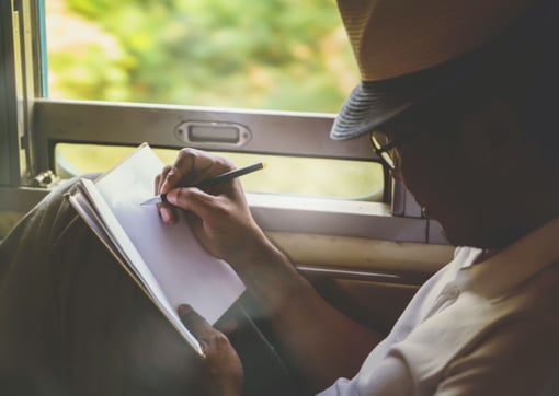 A man in a hat writes in his notebook while he rides on a train.